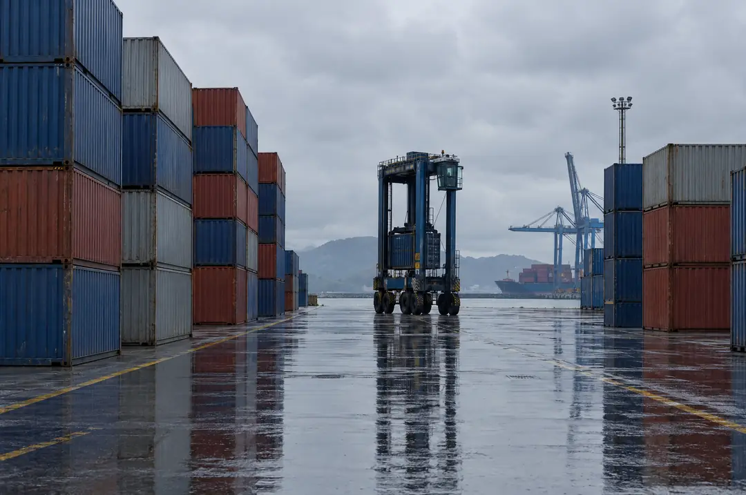 Illustration of a port container yard with stacked shipping containers, a straddle carrier and a vessel at the cranes.